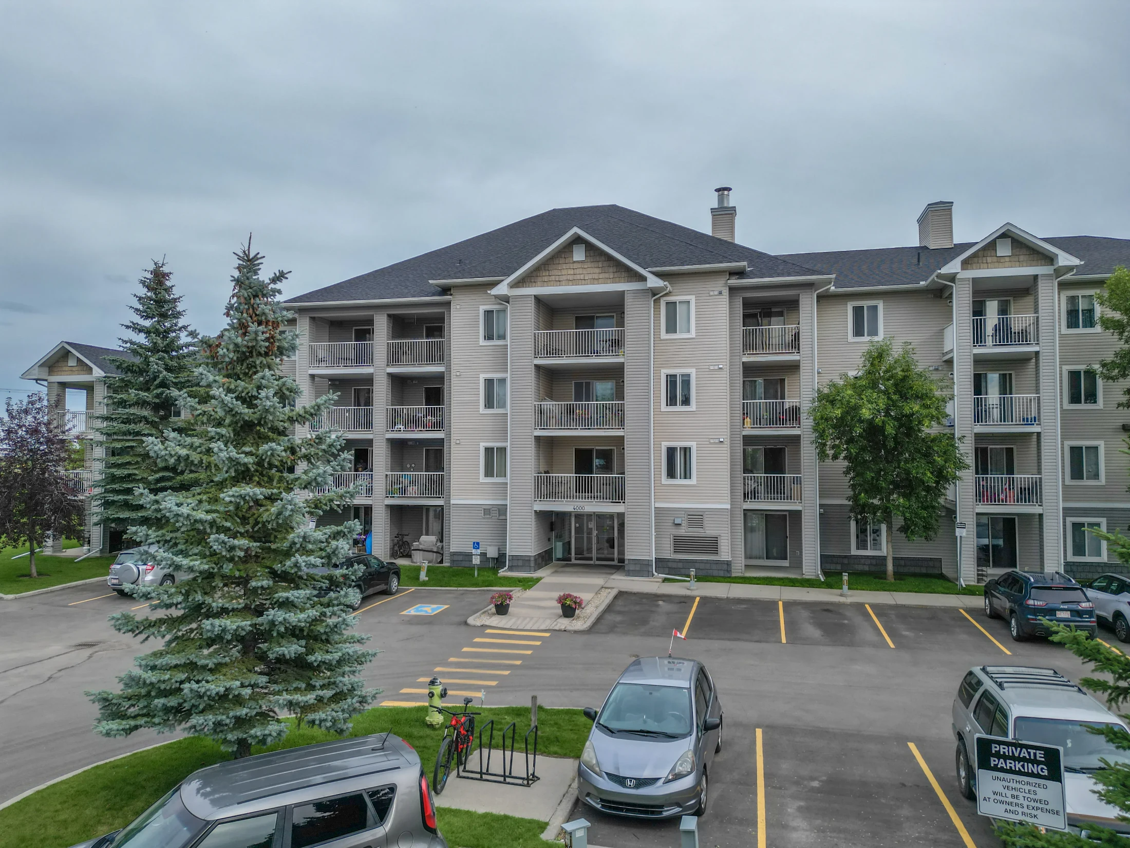 Four-storey condo building with balconies, surface parking and tall spruce trees