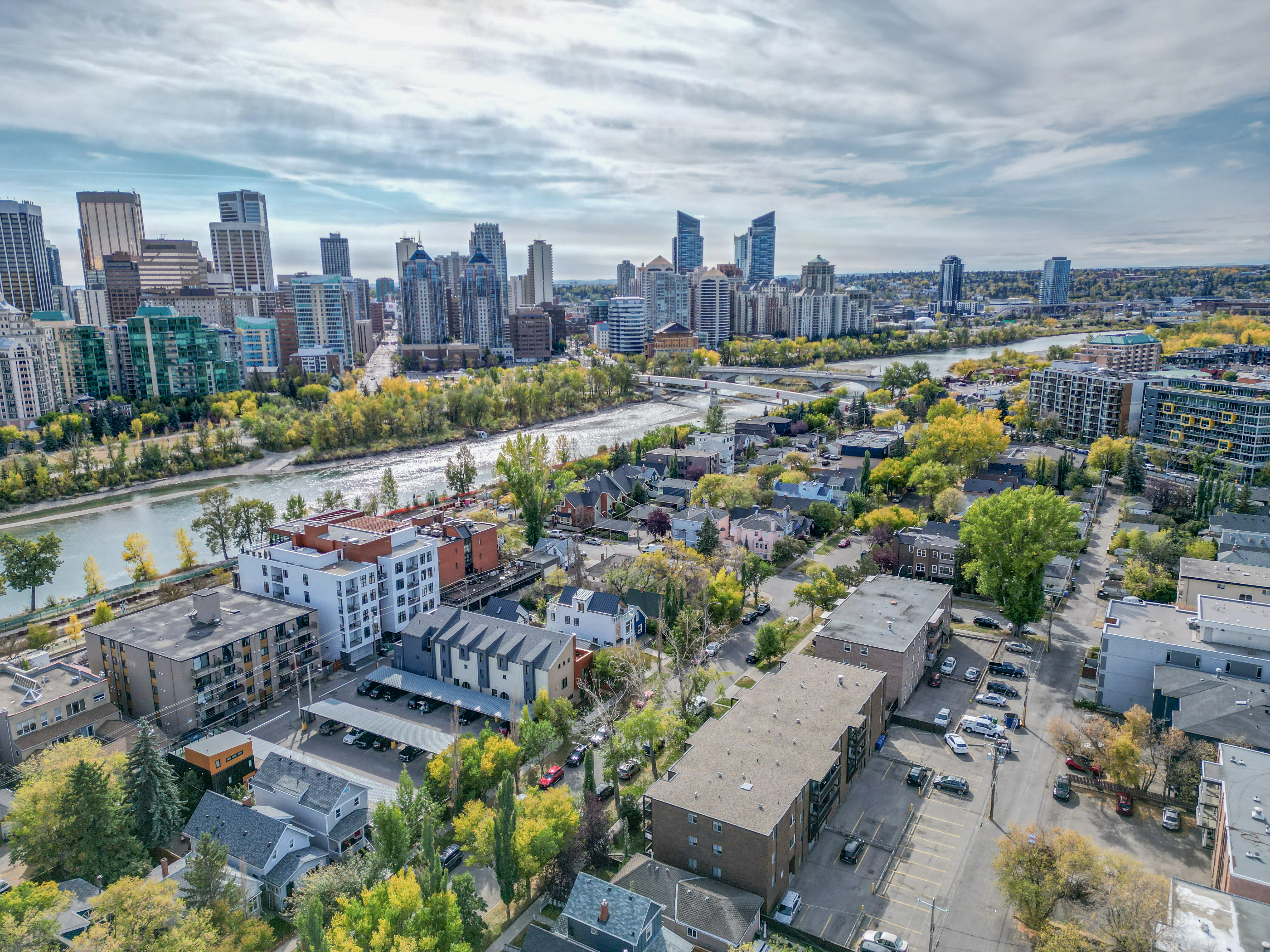 Aerial of inner-city homes and trees along the Bow River with a footbridge and downtown towers
